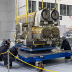 A group of technicians wearing clean room garments, including blue lab coats, hair nets, gloves, and safety glasses, carefully guide a large piece of hardware onto a blue platform inside a high bay facility.
