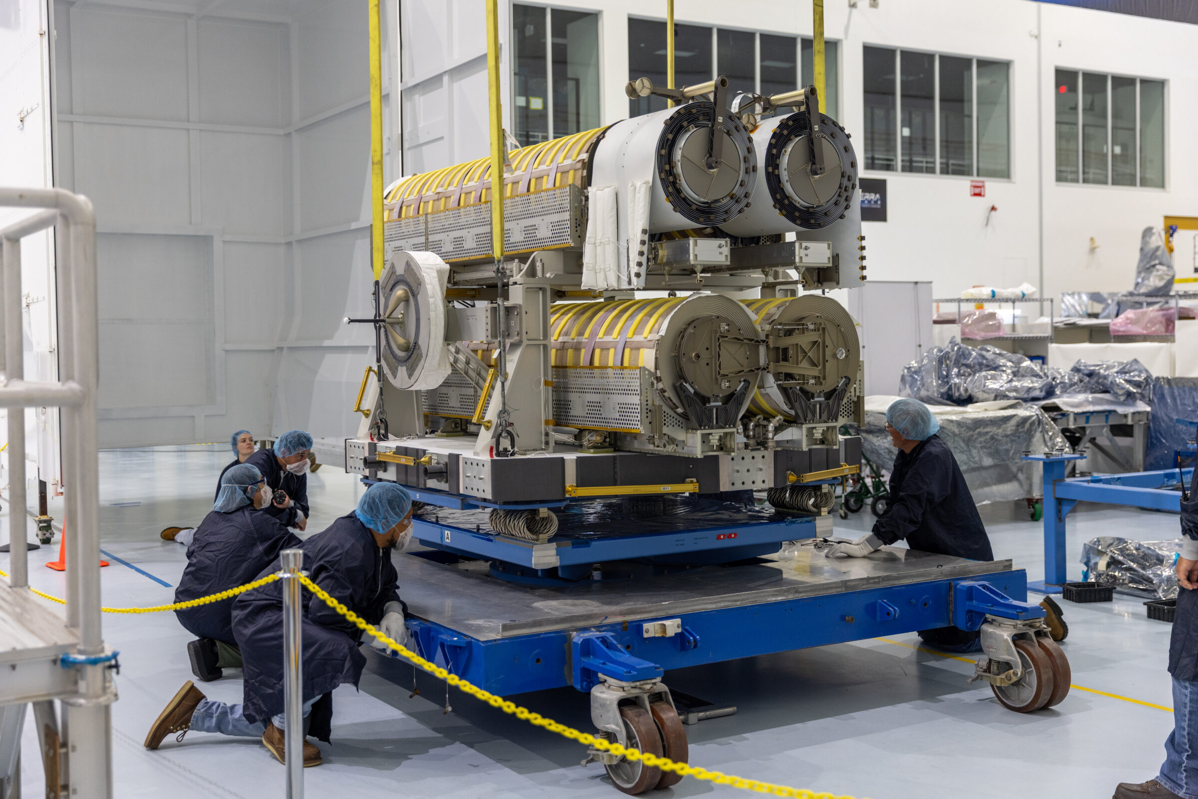 A group of technicians wearing clean room garments, including blue lab coats, hair nets, gloves, and safety glasses, carefully guide a large piece of hardware onto a blue platform inside a high bay facility.