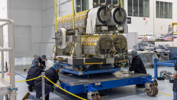 A group of technicians wearing clean room garments, including blue lab coats, hair nets, gloves, and safety glasses, carefully guide a large piece of hardware onto a blue platform inside a high bay facility.